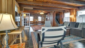 A spacious living room with a high ceiling, wooden beams, a sofa, a chair, a table, and a lamp. - Historic Cottage w/ Beautiful Oceanfront View (Marshfield)