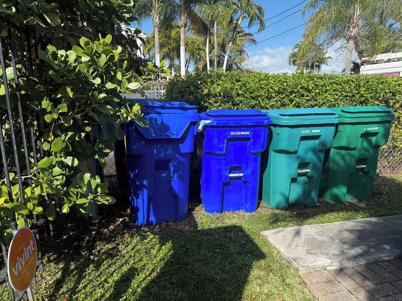 Tres grans dipòsits blaus i verds per a reciclar alineats en una zona verda, amb una haieja i una tanca al fons.