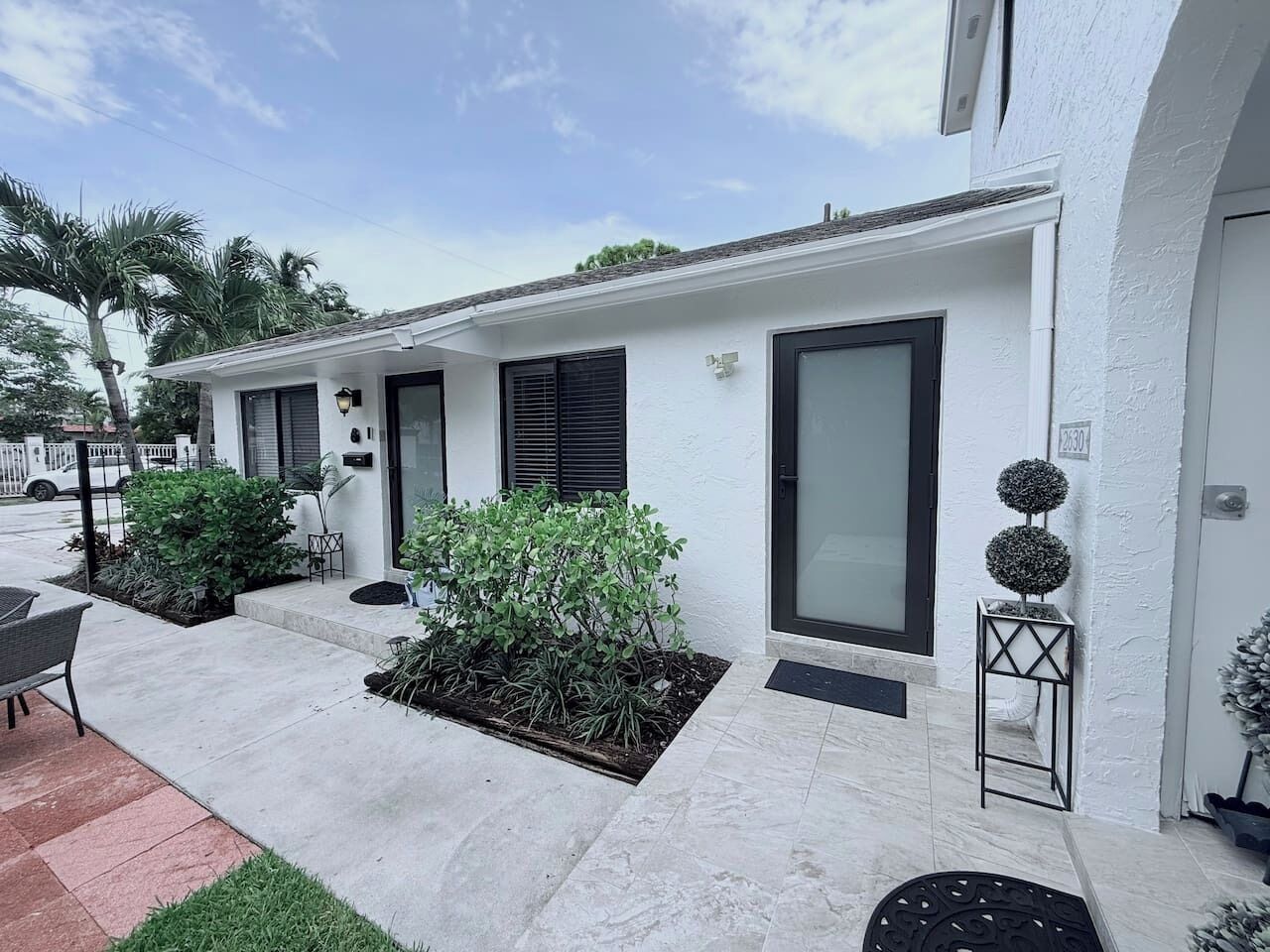 A modern building with a white exterior, black shutters, and a small garden area with plants and a chair.