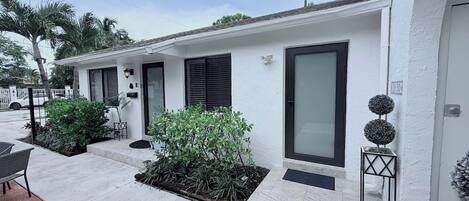 A modern building with a white exterior, black shutters, and a small garden area with plants and a chair.