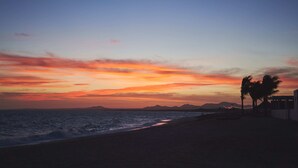 Ein Strand bei Sonnenuntergang mit Palmen und einem kleinen Gebäude.