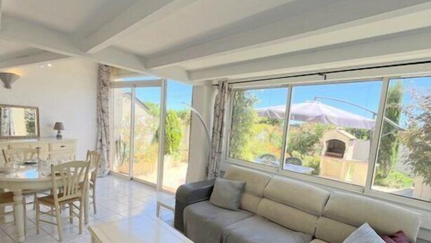 Ein geräumiger Wohnbereich mit Sofa, Couchtisch und Esstisch mit Stühlen. Große Fenster bieten Blick ins Freie.
