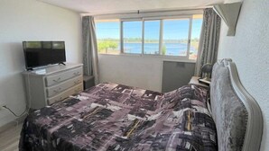 Ein Schlafzimmer mit Bett, Kommode, Fernseher und Fenster mit Blick auf ein Gewässer.
