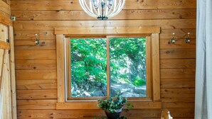 A wooden cabin interior with a wooden table and chairs, a window with a view of greenery, and a hanging light fixture.