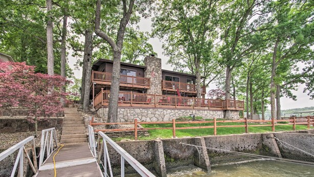 Un pont en bois mène à un bâtiment en pierre doté de balcons, le tout étant entouré d’arbres et agrémenté d’un petit élément décoratif aquatique.