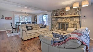 Un vaste salon agrémenté d’une cheminée en pierre, un coin repas avec une table et des chaises, et un espace salon confortable avec un canapé et un pouf.
