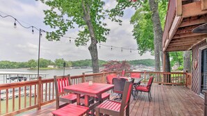 Une terrasse en bois avec des chaises et des tables rouges, donnant sur un lac où l’on peut voir des bateaux et des arbres.