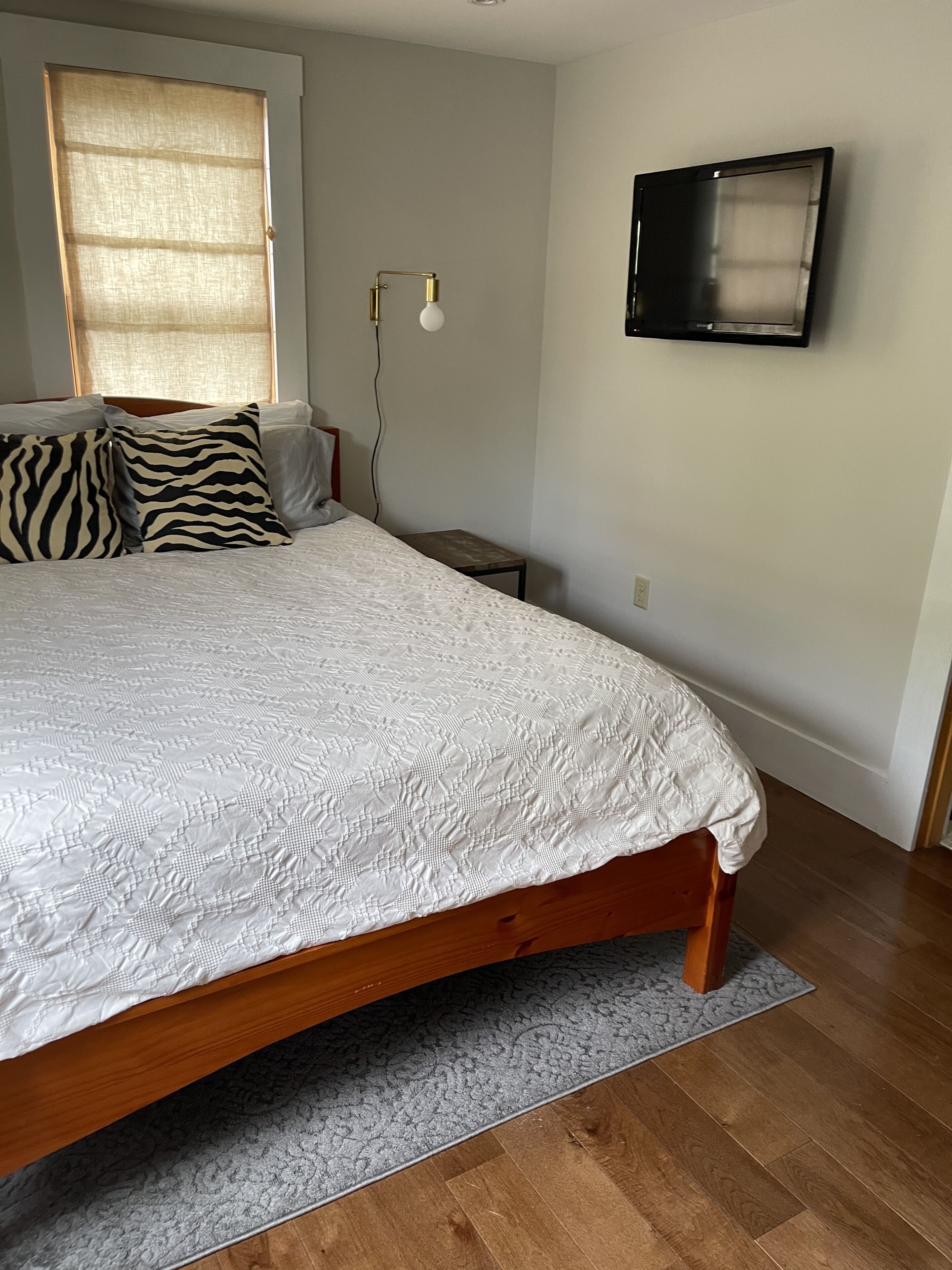 A bedroom with a large bed, a window with blinds, a bedside table, and a wall-mounted TV.
