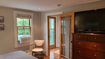 A bedroom with a bed, a chair, a TV on a wooden cabinet, and a window with a view of greenery.