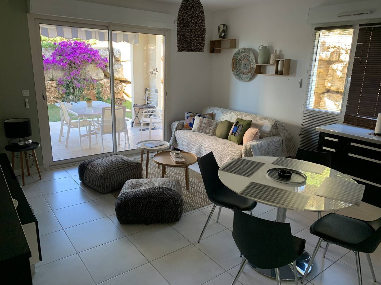 Een moderne woonkamer met een bank, poufs, een eettafel en stoelen. Er is een schuifdeur die leidt naar een buitenruimte met een tafel en stoelen.