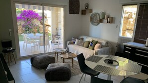 Ein modernes Wohnzimmer mit einer Couch, Hocker, einem Esstisch und Stühlen. Ein großes Schiebeglasfenster führt zu einem Außenbereich mit Tisch und Stühlen.