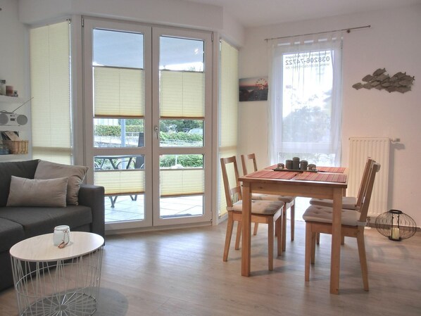 A living room with a sofa, a dining table with chairs, a small round table with a lamp, and large windows with blinds.