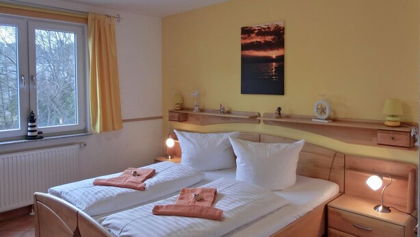 A hotel room with a bed, bedside tables, a window with curtains, a wall-mounted shelf with decorative items, and a painting of a sunset.