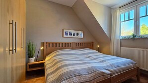 A bedroom with a sloped ceiling, a bed with a striped bedspread, a bedside table with a lamp, a framed picture on the wall, and a window with curtains. - Apartment K5 - 3-room apartment for 5 people. on the 2nd floor (Graal-Müritz)