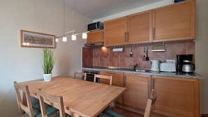 A compact kitchen with wooden cabinets, a microwave, a stove, and a coffee maker. There is a dining table with chairs and a potted plant. - Apartment K5 - 3-room apartment for 5 people. on the 2nd floor (Graal-Müritz)