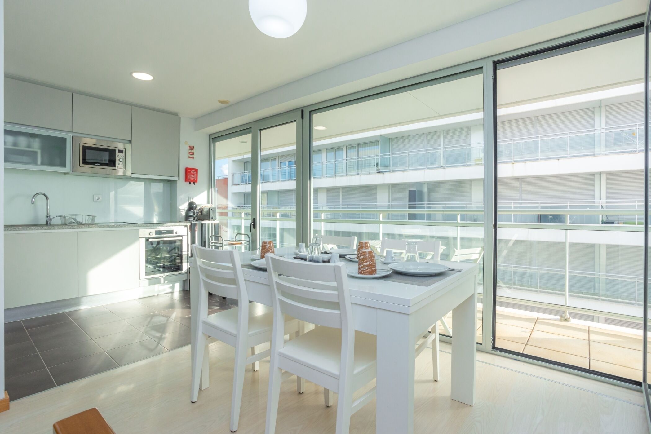 Une cuisine moderne avec des meubles blancs, un four à micro-ondes et un four traditionnel. On y trouve une table à manger avec des chaises et une cloison en verre. La pièce est dotée de grandes fenêtres et offre une vue sur un bâtiment à l’extérieur.