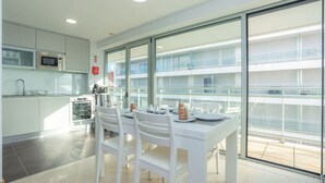A modern kitchen with white cabinets, a microwave, and an oven. There is a dining table with chairs and a glass partition. The room has large windows and a view of a building outside. - Galante Beach Apartment 2 (Figueira da Foz)