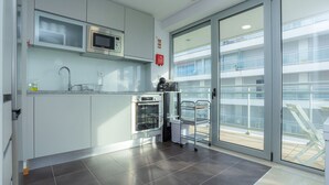 A modern kitchen with a microwave, oven, and sink. There is a sliding glass door leading to a balcony with a view of a building. - Galante Beach Apartment 2 (Figueira da Foz)