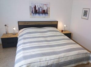 Un lit bien fait, recouvert d’une couvre-lit à rayures, deux tables de chevet avec des lampes, un tableau encadré représentant un paysage urbain accroché au mur, et une petite photo encadrée également accrochée au mur.