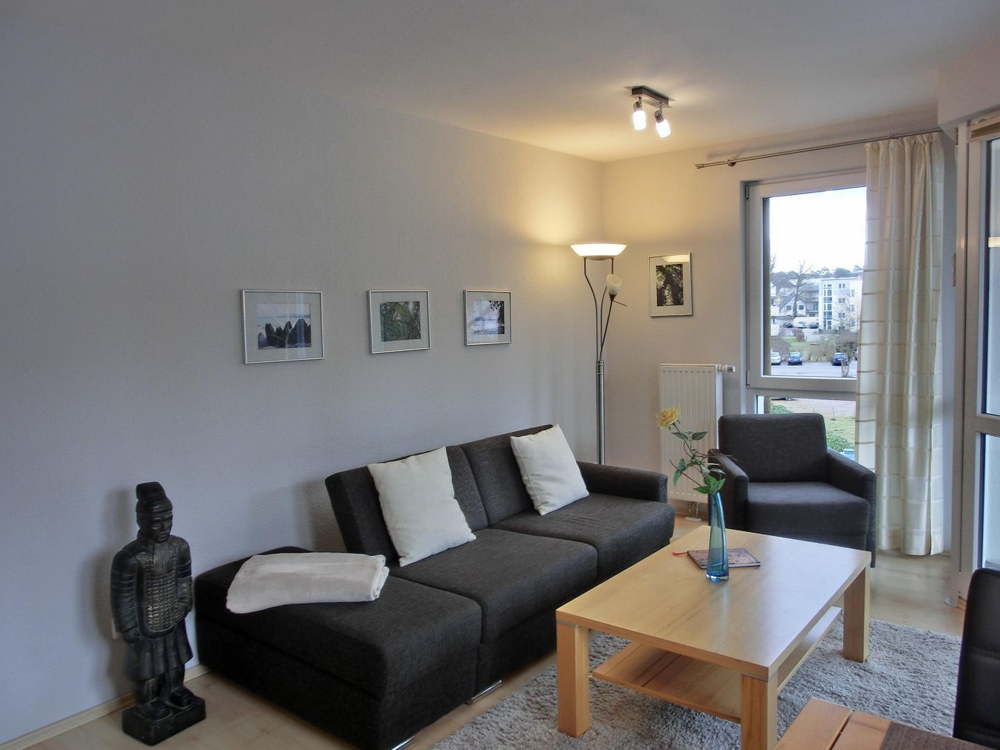 Ein modernes Wohnzimmer mit einem grauen Sofa, einem Holztisch und einem schwarzen Sessel. An der Wand hängen drei gerahmte Bilder und eine Stehlampe.