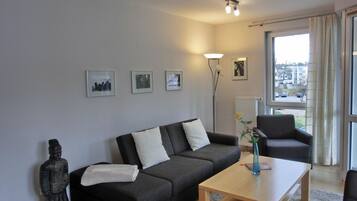 Ein modernes Wohnzimmer mit einem grauen Sofa, einem Holztisch und einem schwarzen Sessel. An der Wand hängen drei gerahmte Bilder und eine Stehlampe.