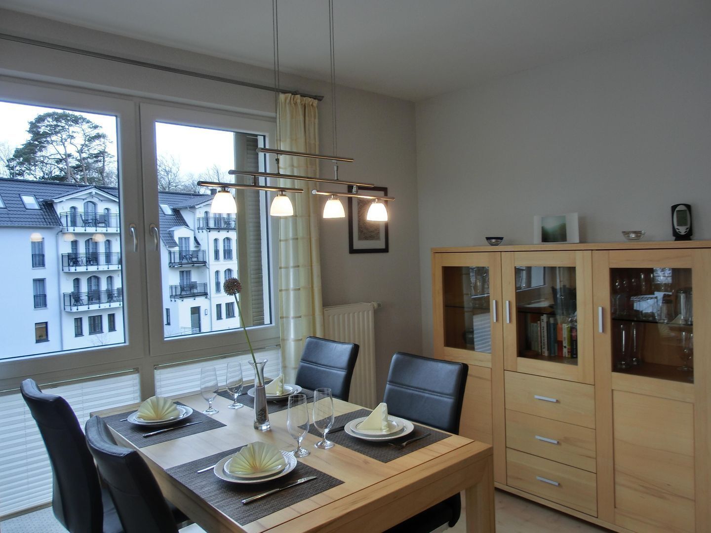 Ein Essbereich mit einem Holztisch, gedeckt für zwei Personen, einem Schrank mit Glastüren und Blick auf Gebäude durch große Fenster.