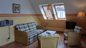Ein gemütliches Wohnzimmer mit einer Couch, zwei Korbstühlen, einem Couchtisch und einem Fernseher. Zwei Fenster mit Vorhängen, ein Deckenventilator und ein gerahmtes Bild sind an den Wänden zu sehen.