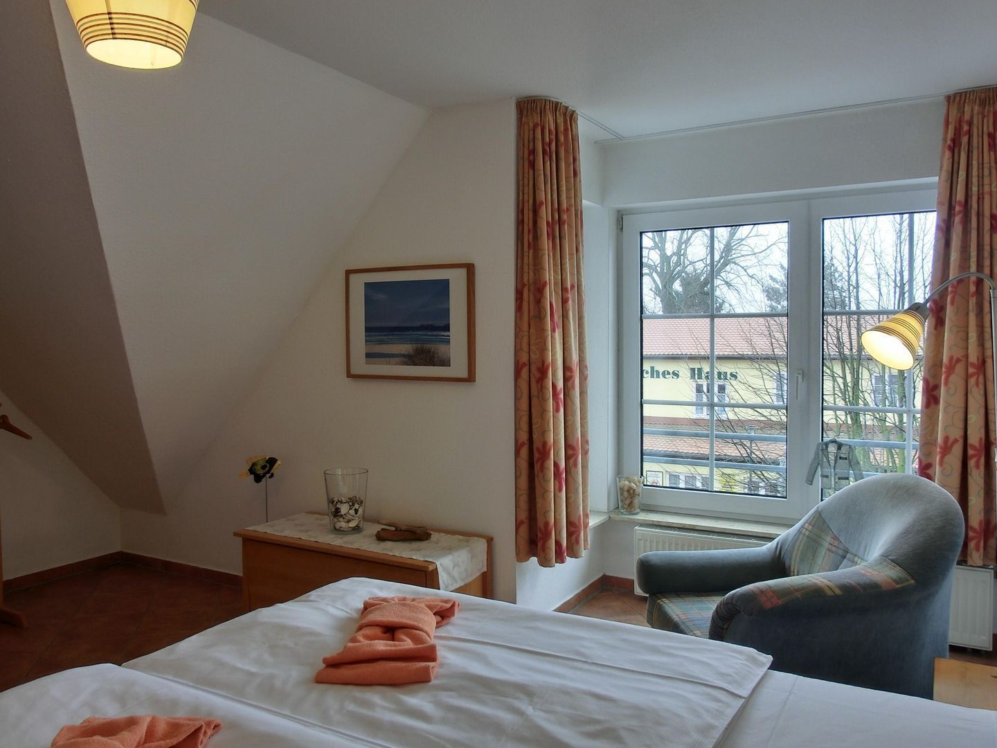 A hotel room with a bed, a chair, a small table with a glass and a vase, a picture on the wall, and a window with curtains.