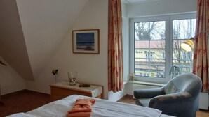 Ein Hotelzimmer mit einem Bett, einem Sessel, einem kleinen Tisch mit einem Glas und einer Vase, einem Bild an der Wand und einem Fenster mit Vorhängen.