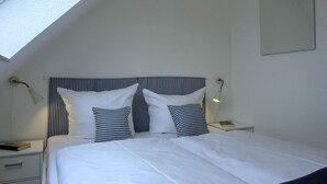 A bedroom with a sloped ceiling, a bed with white bedding and striped pillows, a bedside table with a lamp, and a nightstand with a phone. - Apartment H 07 - Apartment near the beach for 4 + 1 people, with WLAN (Graal-Müritz)