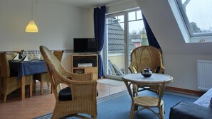A room with a dining area, two wicker chairs, a small table with a teapot, a television on a cabinet, and a large window with a view of the outside. - Apartment H 07 - Apartment near the beach for 4 + 1 people, with WLAN (Graal-Müritz)