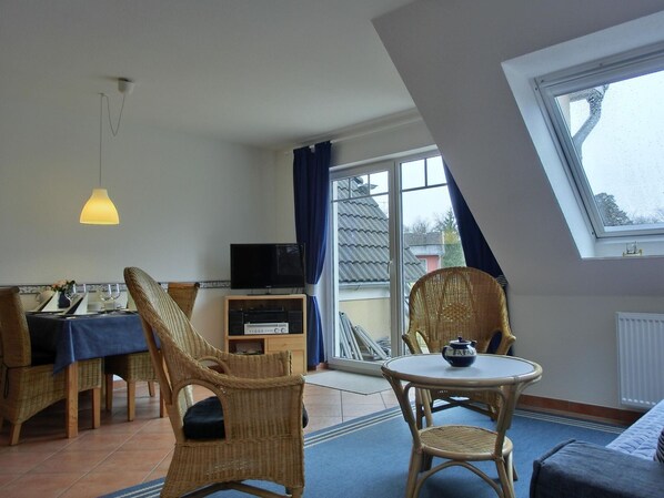 A room with a dining area, two wicker chairs, a small table with a teapot, a television on a cabinet, and a large window with a view of the outside.