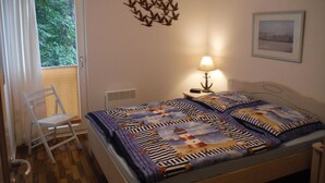 Ein Schlafzimmer mit Bett, Sessel, Nachttisch mit Lampe, Wanddeko und einem Fenster mit Vorhängen.