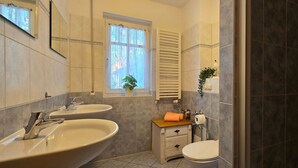A bathroom with two sinks, a toilet, a window with curtains, a towel rack, and a shower area.