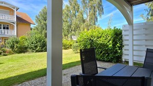 A covered patio with a table and chairs, surrounded by a white fence and lush greenery.