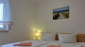 Una cama doble con sábanas blancas y dos toallas rosas, una lámpara de noche y un cuadro de una playa en la pared.