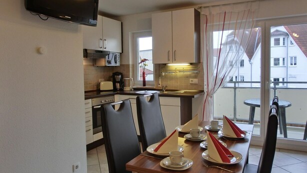 Una cocina moderna con un televisor de pantalla plana, una mesa de comedor para dos personas con servilletas rojas, y vistas a edificios a través de un ventanal grande.