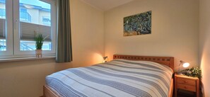 Een slaapkamer met een bed, nachtkastje, lamp, raam met zonwering, een kamerplant en een schilderij aan de muur.
