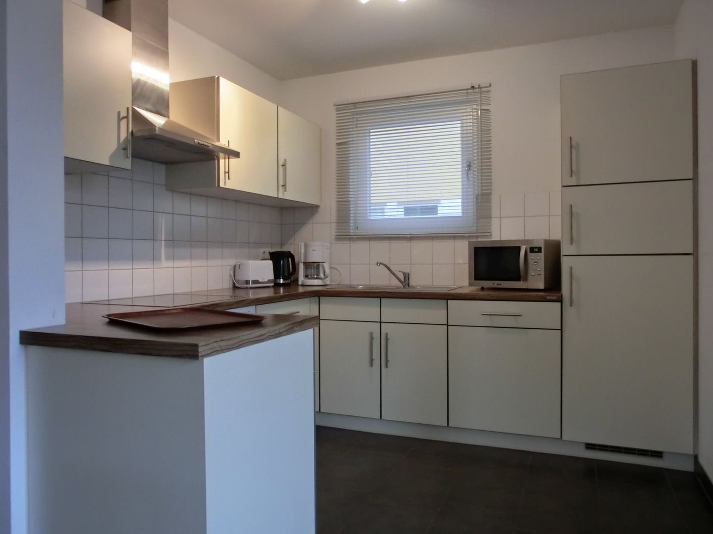 Een moderne keuken met witte kastjes, een magnetron, een koffiezetapparaat en een raam met jaloezieën.