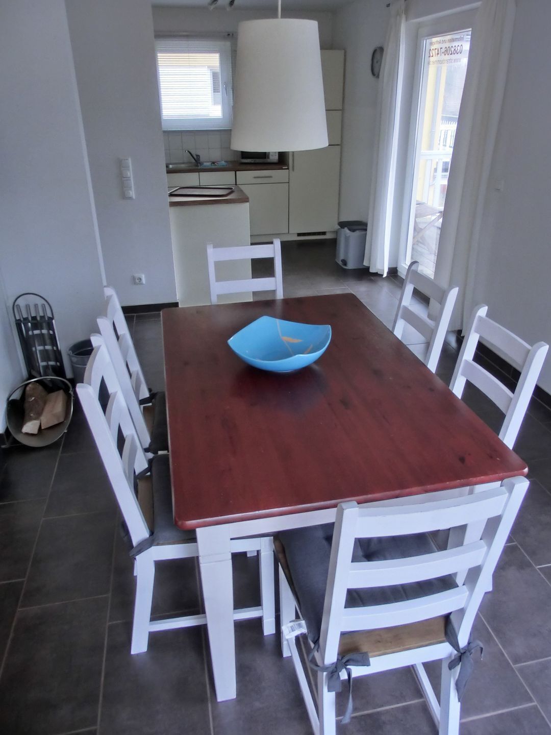 Zona pranzo con tavolo in legno e sedie bianche, una ciotola blu sul tavolo e una cucina sullo sfondo.