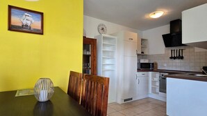 Un comedor con pared amarilla, un cuadro enmarcado y una mesa con sillas. Junto a él, una cocina con armarios blancos, un microondas y un fregadero.