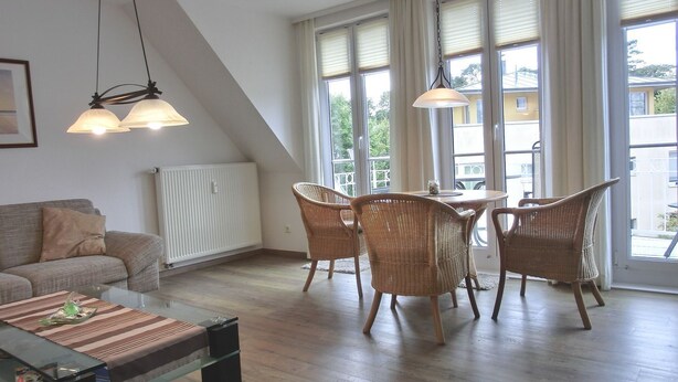 Uma sala de estar moderna com sofá, mesa de centro de vidro, cadeiras de vime e uma mesa de jantar. Grandes janelas oferecem luz natural e vista para o exterior.