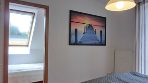 Quarto de hotel com cama, janela, pintura de um cais e uma luminária no teto.