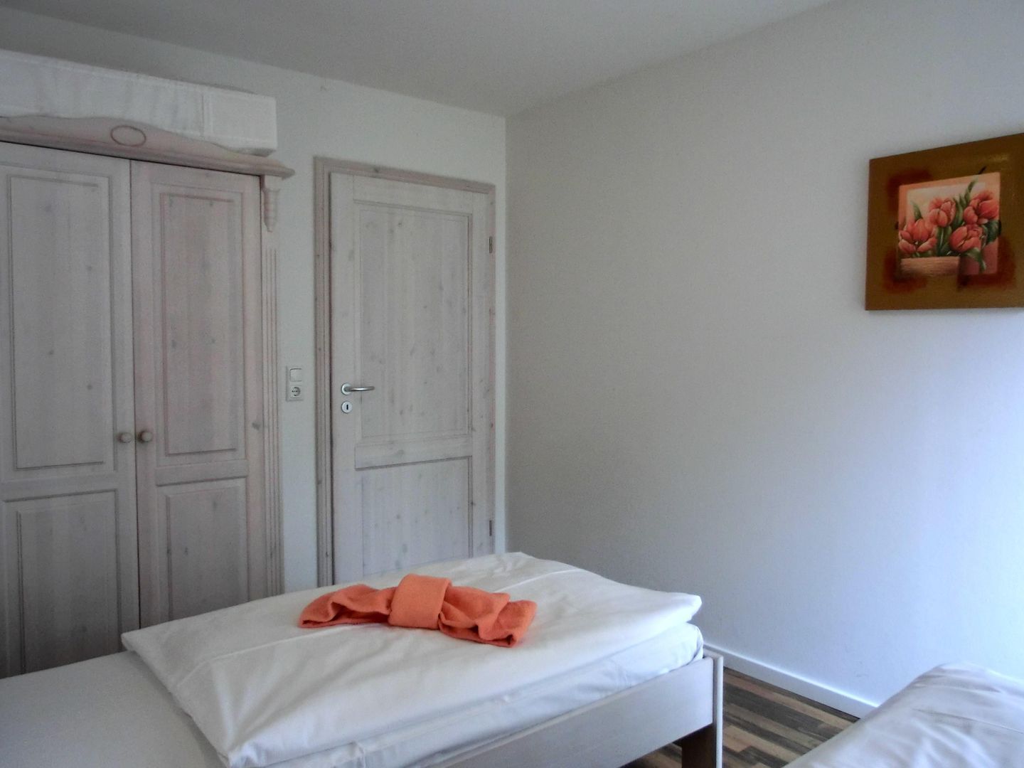 Een kamer met twee witte deuren, een bed met witte lakens en een oranje handdoek, en een bloemen schilderij aan de muur.