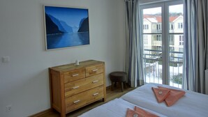 Un dormitorio con una cama, un aparador de madera, un taburete pequeño, una ventana con cortinas y un cuadro de un paisaje.