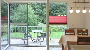Een kamer met een schuifglazen deur naar een balkon met een tafel en stoelen, een eethoek met tafel en stoelen, en een hanglamp.