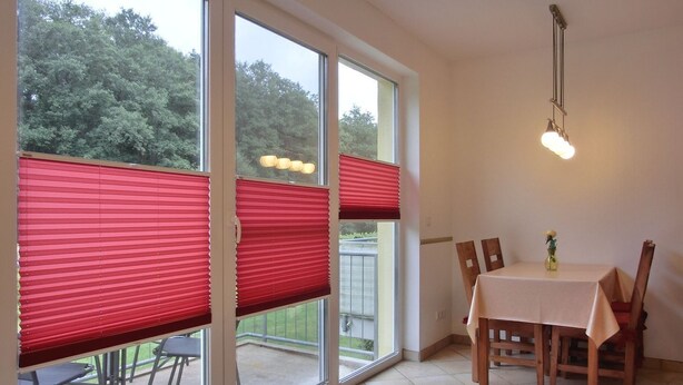 Een eethoek met een tafel en stoelen, grote ramen met rode jaloezieën en een hangende lamp.