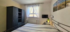 Ein Schlafzimmer mit Bett, Kleiderschrank, einem Fenster mit Vorhängen, einem an der Wand befestigten Fernseher und einem Heizkörper.