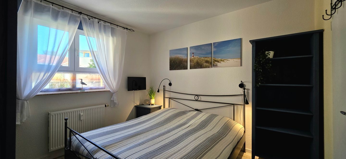 A bedroom with a bed, a window with curtains, a bedside table with a lamp, and wall-mounted artwork.