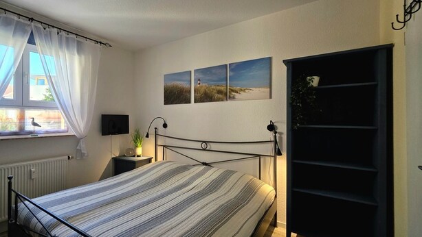 Une chambre à coucher avec un lit, une fenêtre avec des rideaux, une table de chevet avec une lampe et des œuvres d’art accrochées au mur.